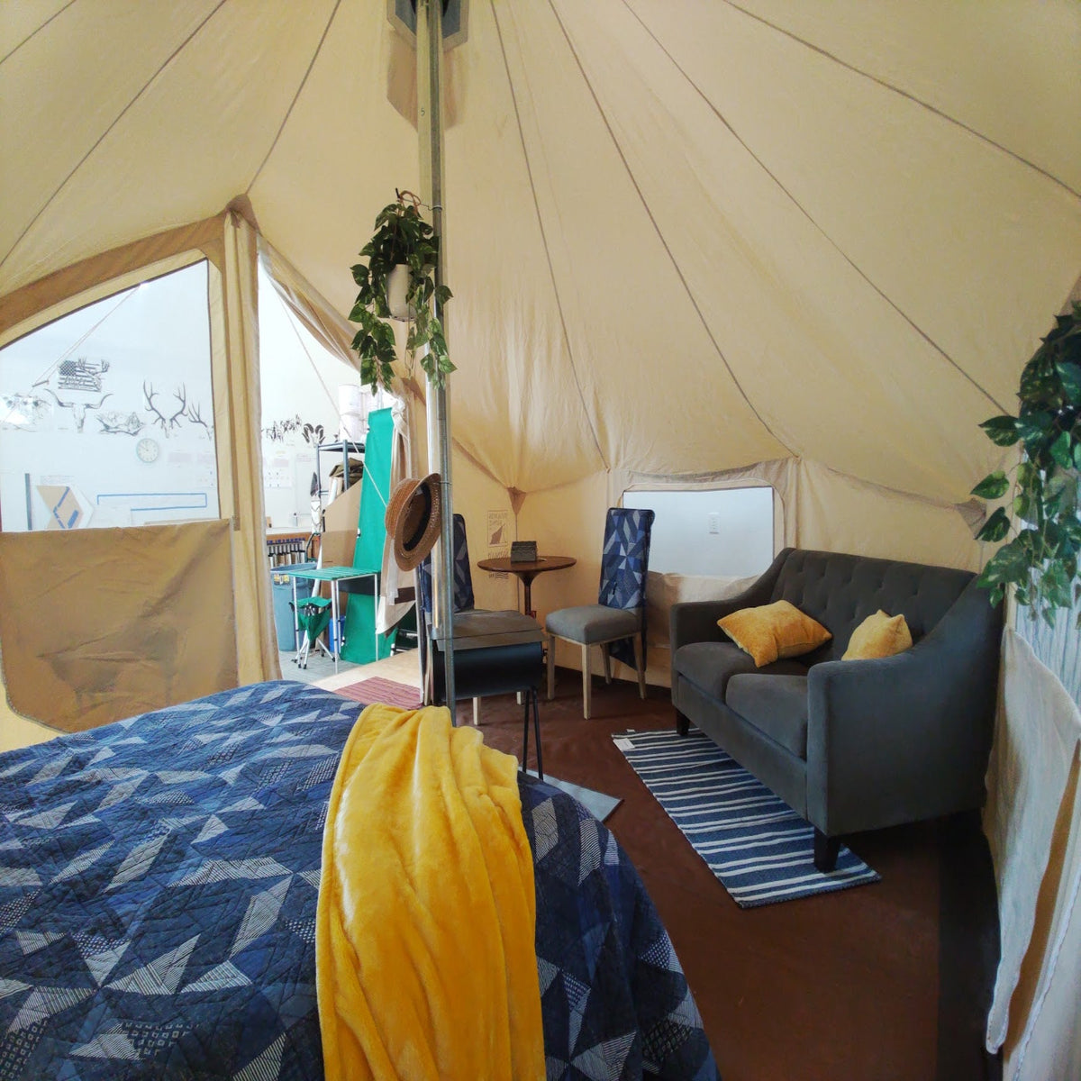 A cozy interior scene inside a large canvas glamping tent, showing a bed with a blue geometric patterned cover and yellow blanket, a gray sofa with yellow pillows, a small dining table, and plants hanging from the tent ceiling.