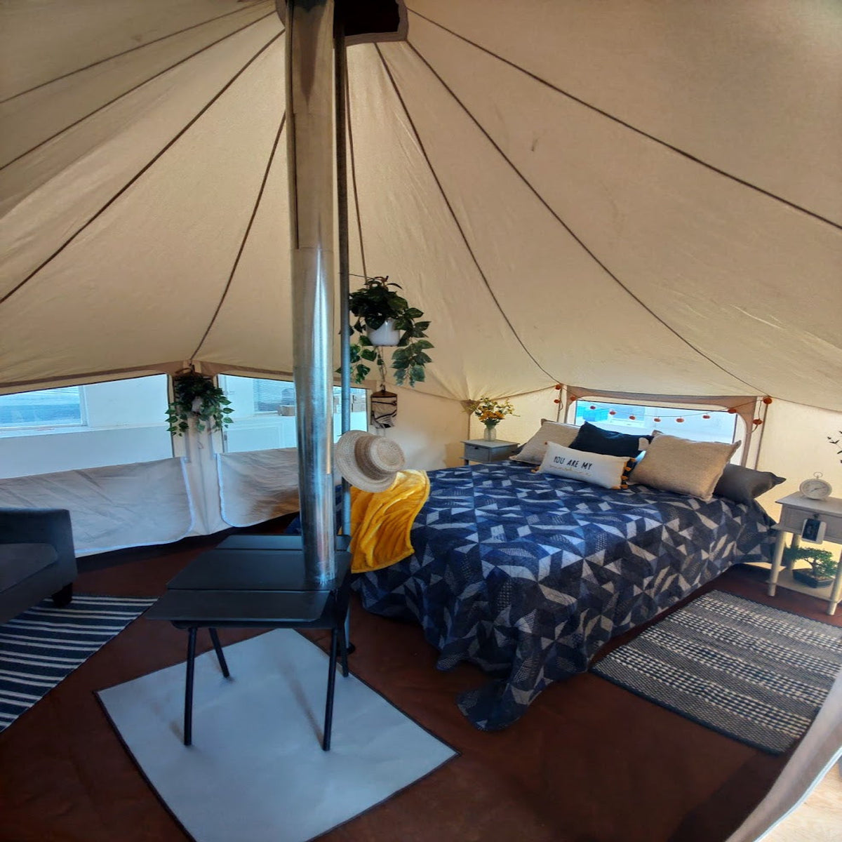 A cozy interior shot of a large canvas glamping tent, showing a made bed with a blue and white patterned duvet, a small black side table, a hanging plant, and a metal chimney pipe against the tent walls.