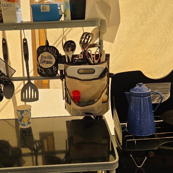 Cream canvas Roamin Home Deluxe Pocket holding a cutting board and cooking utensils, hanging from a camp kitchen shelf beside a stove with a blue coffee pot.