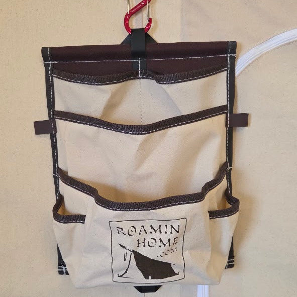 Cream canvas hanging organizer with brown trim, multiple storage pockets, and a Roamin Home logo, suspended from a red carabiner inside a tent.