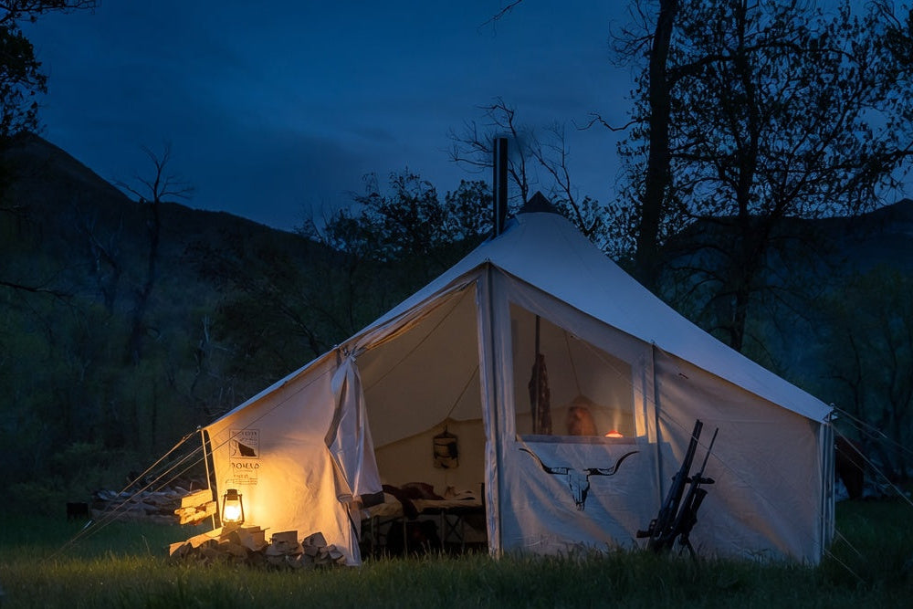 A large white canvas roaminhome tent pitched outdoors at night in a grassy clearing, with warm lights glowing from within and dark trees and mountains in the background.
