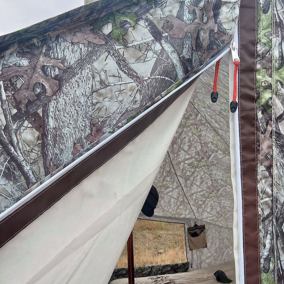 A close-up view of a camouflage tent or shelter with a woodland tree-bark print, cream fabric panels, brown trim, and red drawcord ties. A mirror and small hanging pouch are visible inside the shelter.