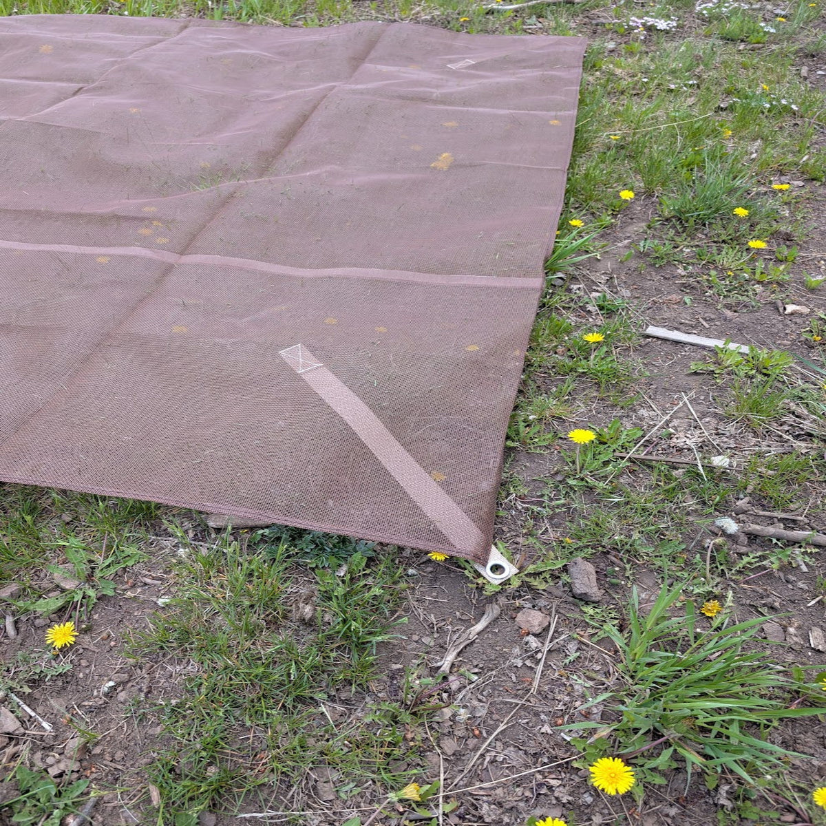 A brown rectangular camping mesh floor laid out on grassy ground, with a metal grommet and strap attached at the corners. Small yellow flowers and dirt are visible around it.