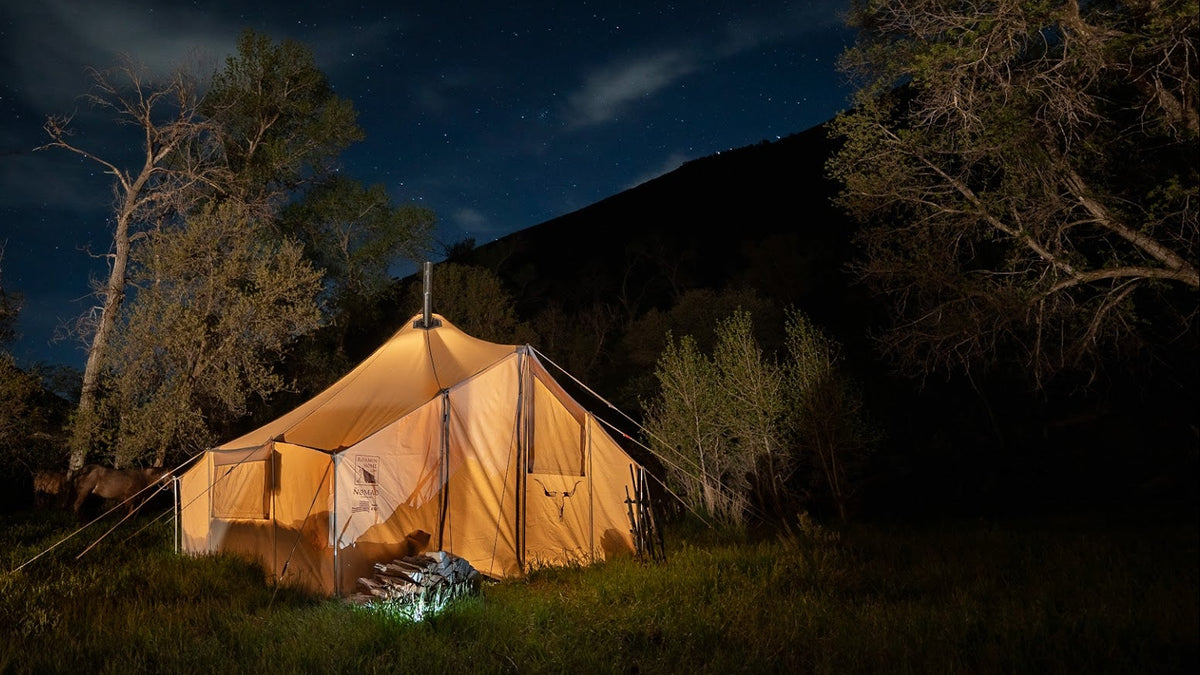 A large canvas camping tent illuminated from within, set up in a grassy field at night under a starry sky with trees and a dark hillside in the background.