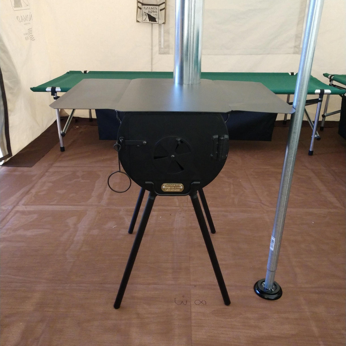 A black metal camp stove with a flat top and four adjustable legs, shown with a silver chimney pipe extending upward. The stove is photographed inside a tent with a brown floor and green camping beds in the background.