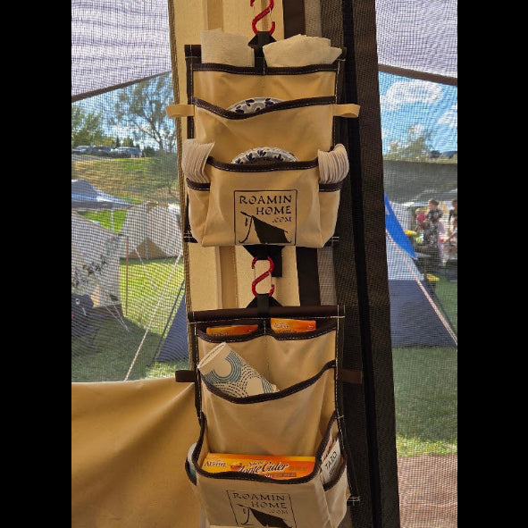 Two cream canvas Roamin Home Deluxe Pockets with brown trim hanging one above the other beside a tent’s mesh doorway, holding plates and other camp essentials.