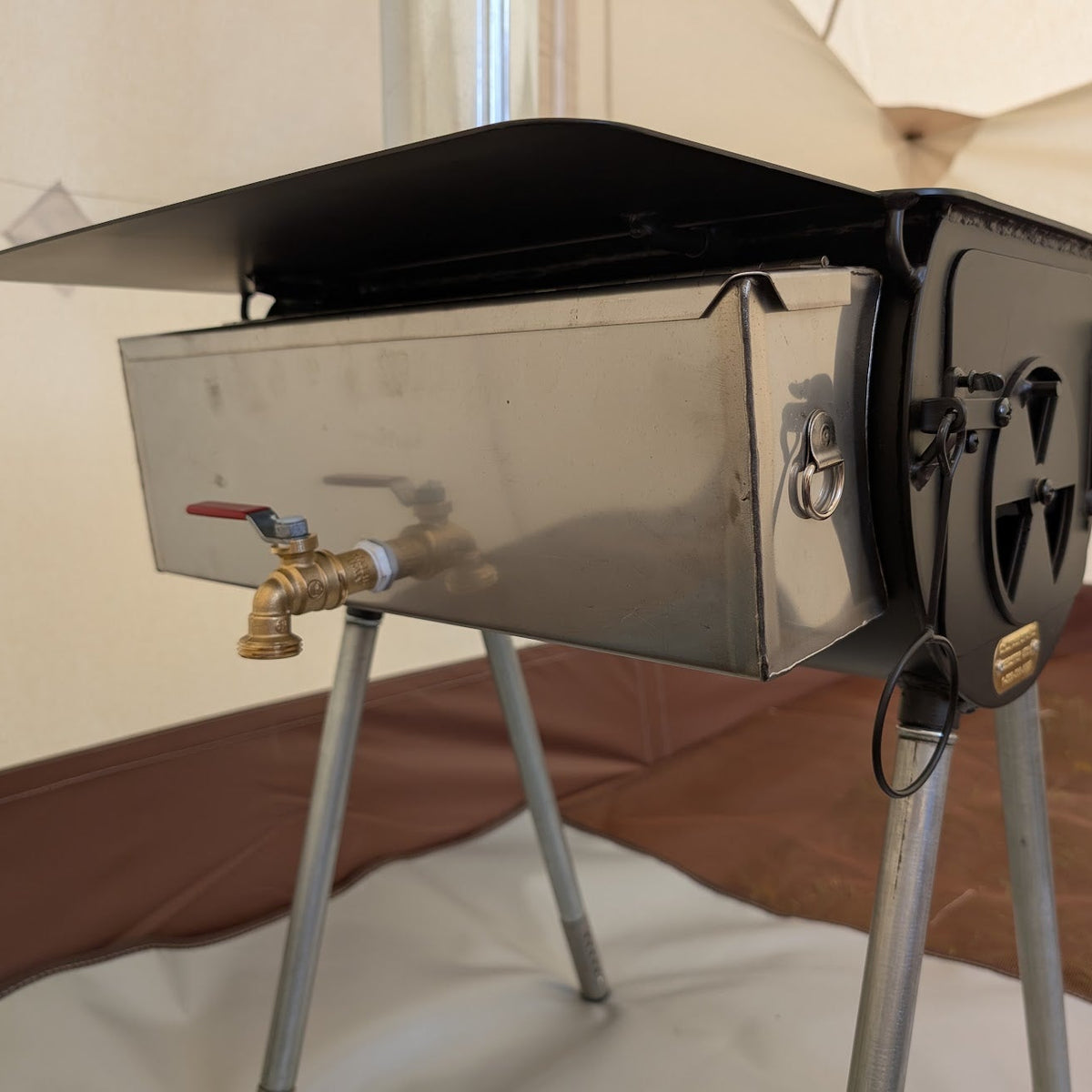 Metal water heater with a brass spigot attached to the side of a wood-burning tent stove inside a canvas tent.