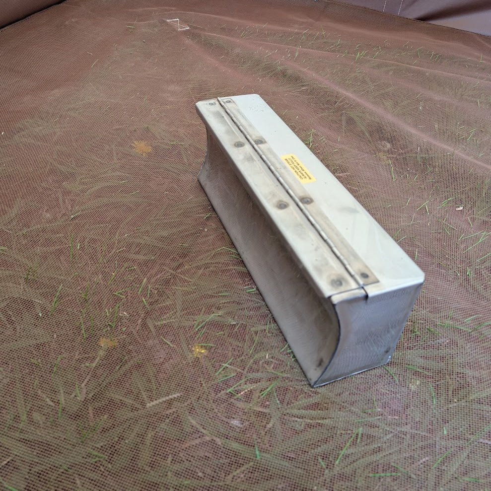 Angled view of a metal tent stove water heater with its hinged lid closed, resting on brown  mesh floor.
