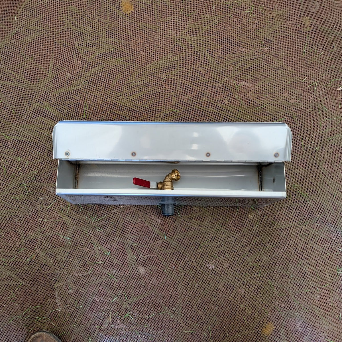 Top view of a metal tent stove water heater with its lid open and a brass spigot with a red handle stored inside.